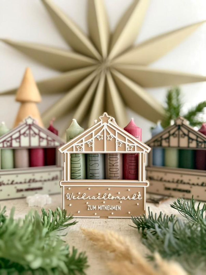 May include: A festive Christmas market-themed display featuring miniature wooden stalls with colorful candles. The stalls are decorated with intricate cut-out designs and the text "Weihnachtsmarkt ZUM MITNEHMEN". A large star-shaped ornament is in the background.