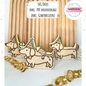 Può includere: Statuine di bassotti in legno con cappelli da festa e papillon. Le statuine sono tagliate al laser e hanno un colore marrone chiaro. L'immagine include anche testo in tedesco.