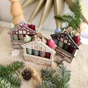 May include: Three miniature wooden advent houses with colorful candles. Each house has a decorative roof and holds four candles in shades of red, pink, green, and blue. The houses are arranged on a neutral-colored surface with pine needles and a pine cone.