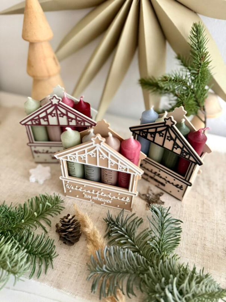 May include: Three miniature wooden advent houses with colorful candles. Each house has a decorative roof and holds four candles in shades of red, pink, green, and blue. The houses are arranged on a neutral-colored surface with pine needles and a pine cone.