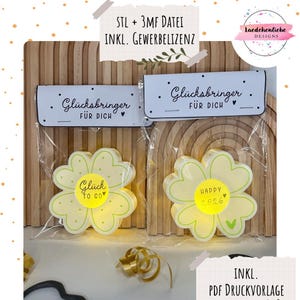 May include: Two flower-shaped LED lights in clear plastic packaging. The lights are yellow with green outlines and have the words "Glück To Go" and "Happy 2026" on them. The packaging includes the text "Glücksbringer FÜR DICH".
