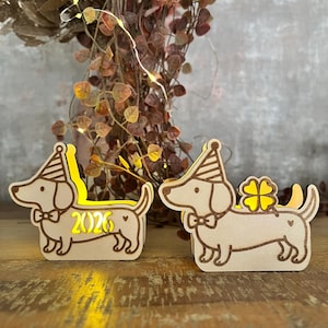 May include: Two wooden dachshund dog figurines with party hats and bow ties. One has the number "2026" illuminated, the other has a four-leaf clover. The dogs are on a wooden surface, with decorative foliage in the background.