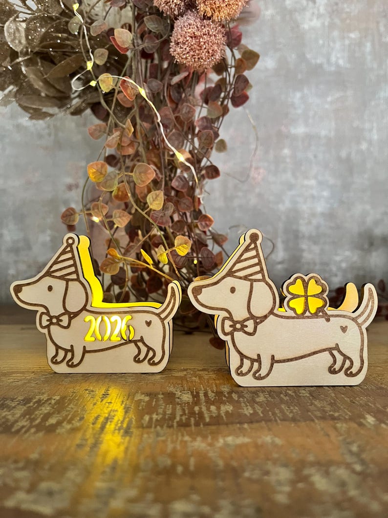 May include: Two wooden dachshund dog figurines with party hats and bow ties. One has the number "2026" illuminated, the other has a four-leaf clover. The dogs are on a wooden surface, with decorative foliage in the background.
