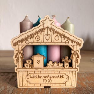 May include: A laser-cut wooden stand shaped like a market stall, featuring engraved holiday decorations and the text "Weihnachtsmarkt TO GO". Four taper candles in muted shades of taupe, blue, pink, and sage green stand behind the wooden structure.