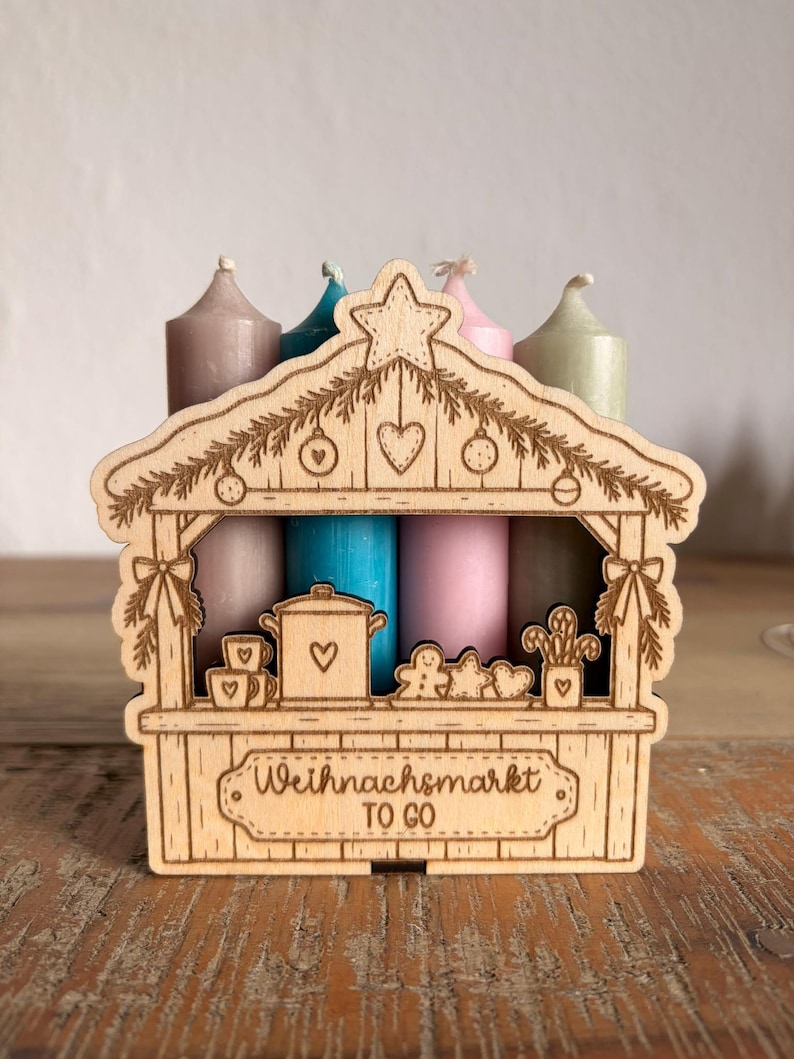 May include: A laser-cut wooden stand shaped like a market stall, featuring engraved holiday decorations and the text "Weihnachtsmarkt TO GO". Four taper candles in muted shades of taupe, blue, pink, and sage green stand behind the wooden structure.