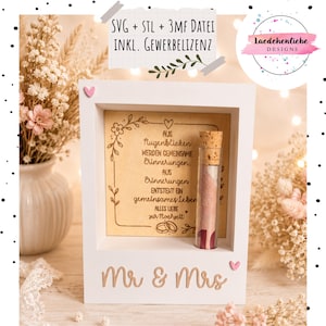 May include: A white wooden shadow box frame featuring the text 'Mr & Mrs' at the base and a central wooden panel engraved with German text and floral motifs. A glass test tube with a cork stopper containing a rolled banknote is attached to the frame.