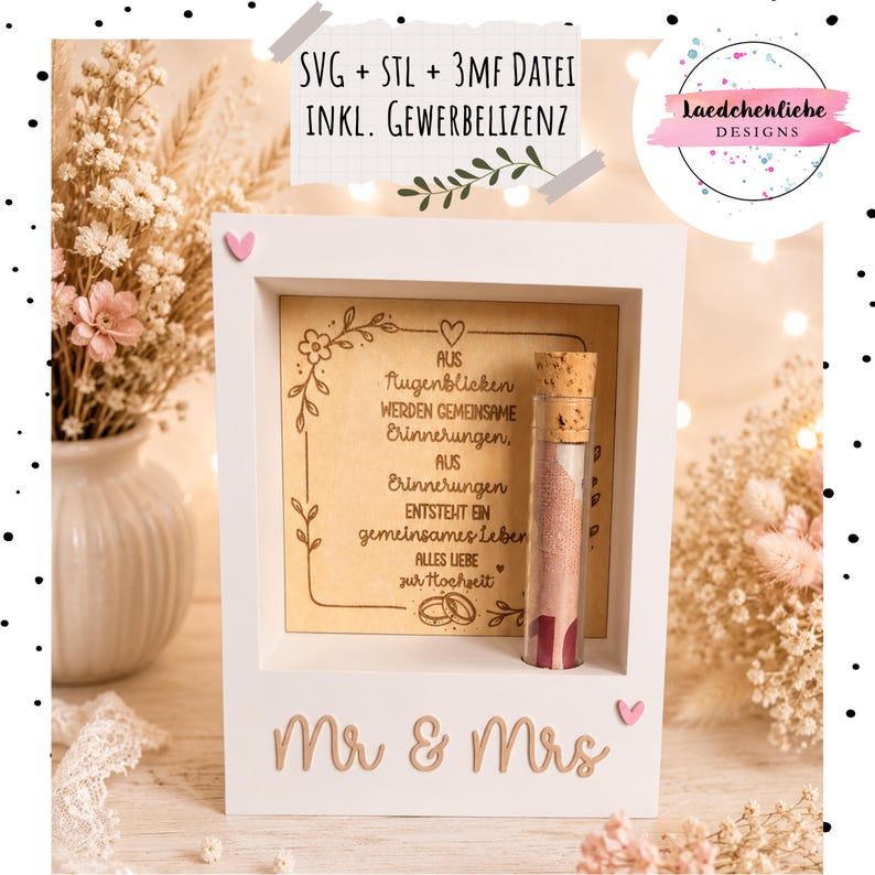 May include: A white wooden shadow box frame featuring the text 'Mr & Mrs' at the base and a central wooden panel engraved with German text and floral motifs. A glass test tube with a cork stopper containing a rolled banknote is attached to the frame.