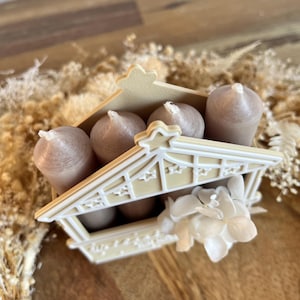 May include: A decorative candle holder shaped like a small house, holding several brown candles. The house is cream-colored with star-shaped cutouts and a floral accent. The candles have white wicks.