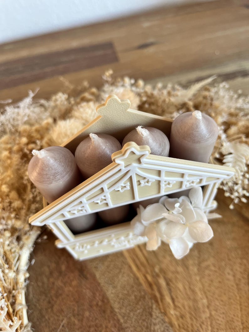 May include: A decorative candle holder shaped like a small house, holding several brown candles. The house is cream-colored with star-shaped cutouts and a floral accent. The candles have white wicks.