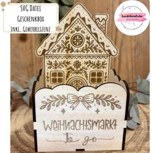 Könnte beinhalten: Eine lasergeschnittene Geschenkbox aus Holz in Form eines Lebkuchenhauses mit aufwendigen Details. Die Box hat einen hausförmigen Aufsatz und einen gewellten Behälter mit dem Text "Weihnachtsmarkt to go". Das Design umfasst eine Schleife und florale Elemente.