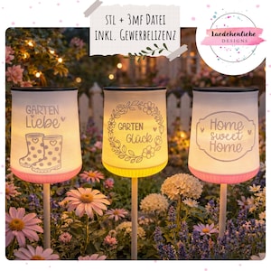 Puede incluir: Tres faroles de jardín iluminados con pantallas blancas y bases rosas o amarillas. Cada farol tiene un diseño y texto diferente: "Garten Liebe", "Garten Glück" y "Home sweet Home". Los faroles están en un jardín.