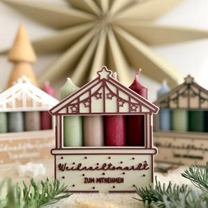 May include: A decorative Christmas market-themed candle holder with four candles in shades of green, pink, and red. The holder is white with a red roof and star accents. The text "Weihnachtsmarkt" is visible.