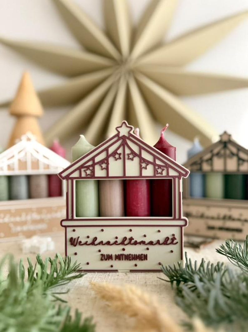 May include: A decorative Christmas market-themed candle holder with four candles in shades of green, pink, and red. The holder is white with a red roof and star accents. The text "Weihnachtsmarkt" is visible.