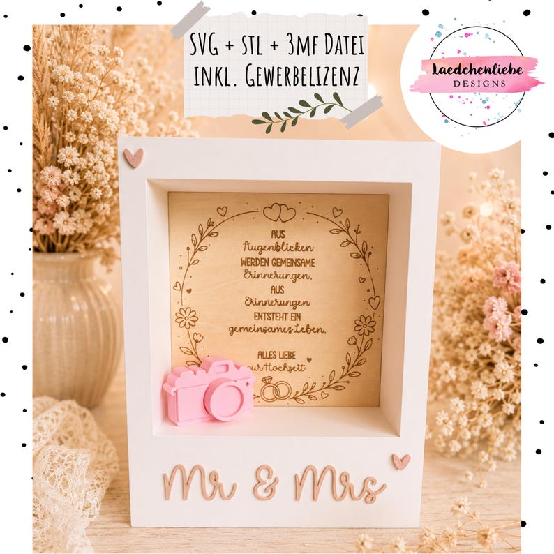 May include: A white wooden shadow box frame featuring a light wood back panel engraved with a floral wreath, two wedding rings, and German text. A small pink 3D camera icon is attached inside, and the words Mr & Mrs are in pink on the front.