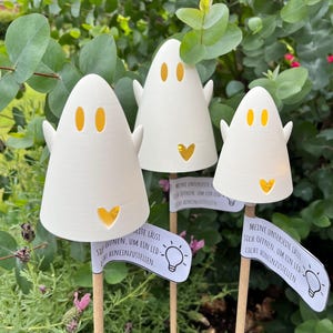 May include: Three white ghost-shaped LED lights with orange eye and heart cutouts. Each ghost has small arms and is mounted on a wooden stick. Small signs with German text and a lightbulb graphic are attached to the sticks.