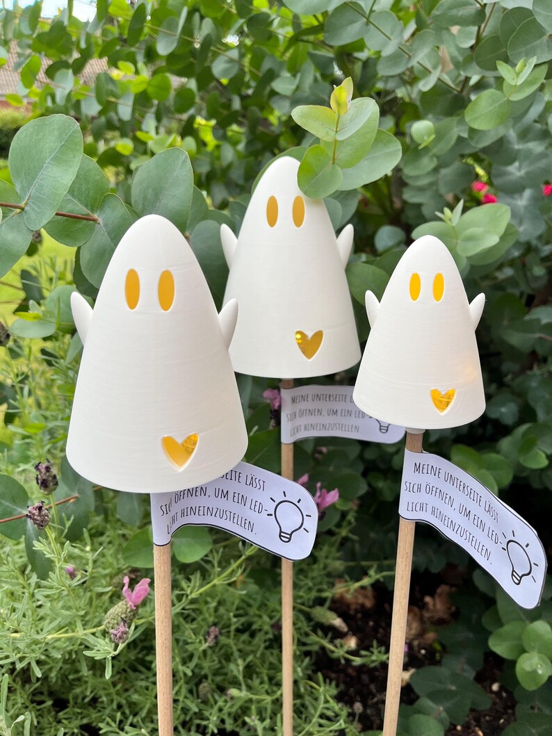 May include: Three white ghost-shaped LED lights with orange eye and heart cutouts. Each ghost has small arms and is mounted on a wooden stick. Small signs with German text and a lightbulb graphic are attached to the sticks.