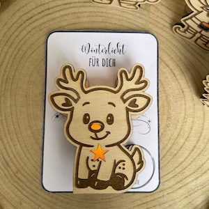 May include: Wooden reindeer Christmas ornament with a star and the text "Winterlicht F&Uuml;R DICH" on a white card. The reindeer has a smiling face, orange nose, and a star in its front legs. Other wooden Christmas ornaments are in the background.