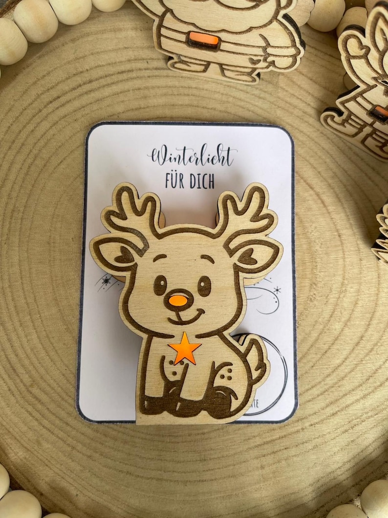 May include: Wooden reindeer Christmas ornament with a star and the text "Winterlicht F&Uuml;R DICH" on a white card. The reindeer has a smiling face, orange nose, and a star in its front legs. Other wooden Christmas ornaments are in the background.