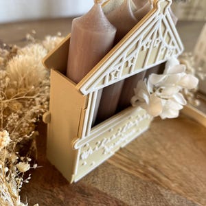 May include: A decorative candle holder in the shape of a house, with a light beige exterior and white accents. The holder contains three beige candles. A white flower is attached to the side of the holder.