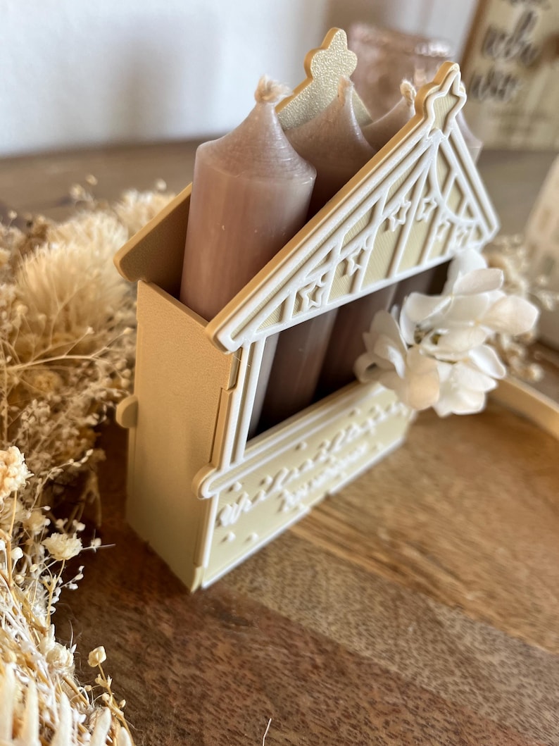 May include: A decorative candle holder in the shape of a house, with a light beige exterior and white accents. The holder contains three beige candles. A white flower is attached to the side of the holder.