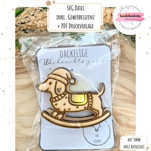 May include: A wooden dachshund Christmas ornament on a rocking horse, wearing a Santa hat and saddle. The ornament is on a white card with German text. The card is in a clear plastic bag.