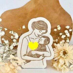 May include: A wooden cutout of a mother holding a child, with a yellow heart in the centre. The cutout is on a light brown surface with white flowers.