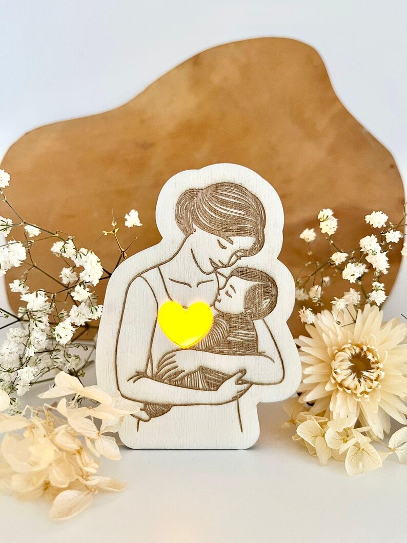 May include: A wooden cutout of a mother holding a child, with a yellow heart in the centre. The cutout is on a light brown surface with white flowers.