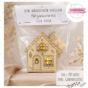 May include: A light-coloured wooden house cutout with engraved details including windows, a door, and a chimney, packaged in a clear plastic bag with a header card that reads EIN HÄUSCHEN VOLLER Herzenswärme FÜR DICH and SVG + PDF DATEI INKL. GEWERBELIZENZ.