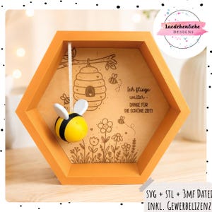 Puede incluir: Un marco hexagonal de madera naranja que contiene un panel de madera clara con una colmena, flores y abejas grabadas con láser. El texto en alemán dice Ich fliege weiter - DANKE FÜR DIE SCHÖNE ZEIT. Una pequeña figura de abeja amarilla y negra cuelga de una cinta.