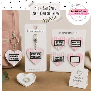 Könnte beinhalten: Herzförmige Rucksackanhänger mit rosa-weißen Streifen und schwarzen Textfeldern mit Aufschriften wie 'Handy ran Mama dran' und 'Notfall Kontakt'. Das Set enthält einen Metallschlüsselring, ein leeres Herz-Tag und eine kleine Infokarte.