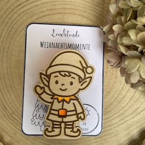 May include: Wooden Christmas elf ornament with a smiling face, wearing a hat and outfit with orange accents. The ornament is on a white card with the text "Leuchtende Weihnachtsmomente". The background is a wooden surface.