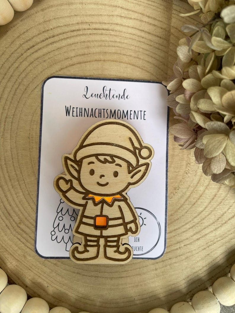 May include: Wooden Christmas elf ornament with a smiling face, wearing a hat and outfit with orange accents. The ornament is on a white card with the text "Leuchtende Weihnachtsmomente". The background is a wooden surface.