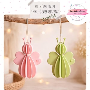 Può includere: Due ornamenti sospesi a forma di fate stilizzate, uno rosa e uno verde, appesi a un ramo. Gli ornamenti hanno un corpo a coste, ali a forma di cuore e antenne. Il testo nell'immagine recita: "STL + 3MF DATEI INKL. GEWERBELIZENZ" e "Laedchenliebe DESIGNS".