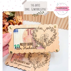 May include: Wooden money holder with a heart-shaped floral design and a couple illustration. The holder has the text "Alles Liebe zur Hochzeit" and is secured with gold-colored heart-shaped studs. A ten-euro bill is visible.