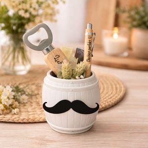 May include: A white ceramic barrel-shaped container with a black mustache design, holding a bottle opener, pen, and decorative elements. The pen has the text "Du bist perfekt". The container sits on a woven mat.