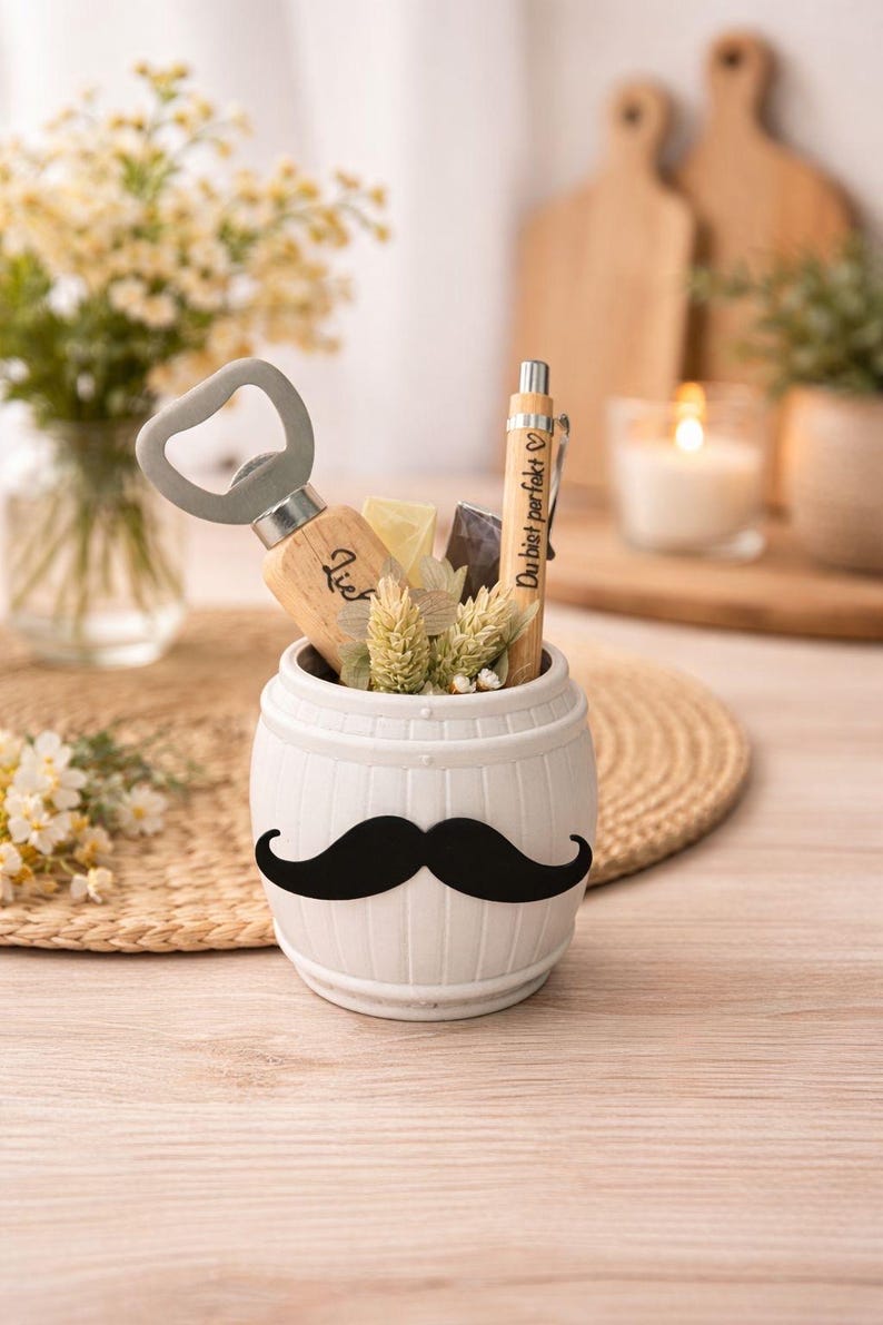 May include: A white ceramic barrel-shaped container with a black mustache design, holding a bottle opener, pen, and decorative elements. The pen has the text "Du bist perfekt". The container sits on a woven mat.