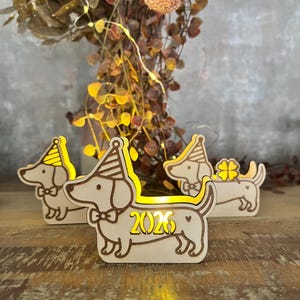 May include: Wooden dachshund dog figurines with party hats and bow ties. The figurines are illuminated with a warm yellow glow, and one displays the year "2026". The decorations are set on a wooden surface.