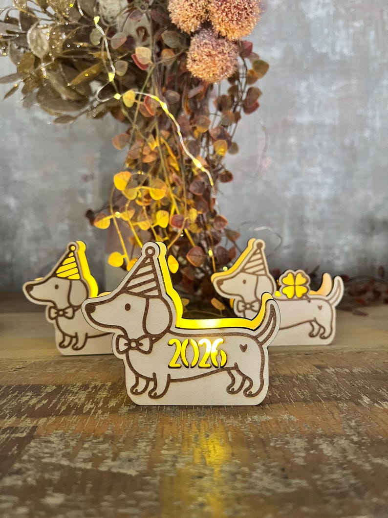 May include: Wooden dachshund dog figurines with party hats and bow ties. The figurines are illuminated with a warm yellow glow, and one displays the year "2026". The decorations are set on a wooden surface.