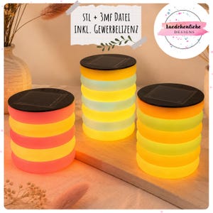 Puede incluir: Tres faroles solares con diseño cilíndrico. Cada farol tiene una parte superior negra y un cuerpo compuesto por bandas horizontales coloridas en tonos amarillos, naranjas y rosas. Los faroles están colocados sobre una superficie de madera, emitiendo un brillo cálido.