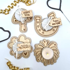 May include: A collection of wooden candle holders and decorations for the New Year. The set includes a number 4, a horseshoe, a flower, and a firework design, each with a tea light candle. The decorations have the phrases "Happy 2026" and "Happy New Year".