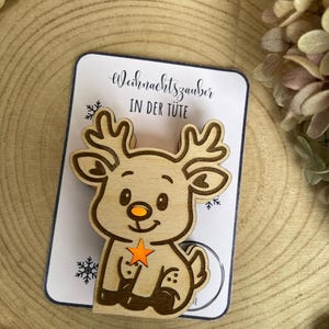 May include: Wooden reindeer ornament with an orange star and nose, sitting on a card that reads "Weihnachtszauber IN DER T&Uuml;TE". The reindeer has detailed antlers and is set against a wooden background, perfect for Christmas.