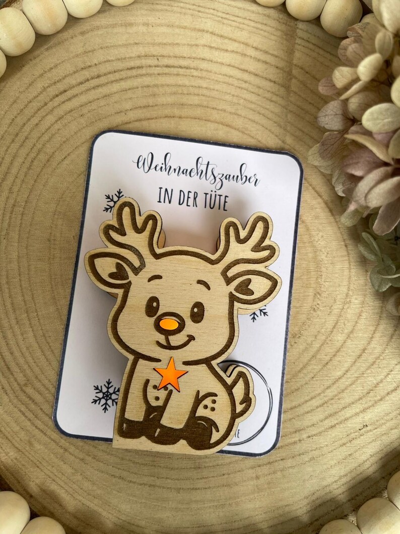 May include: Wooden reindeer ornament with an orange star and nose, sitting on a card that reads "Weihnachtszauber IN DER T&Uuml;TE". The reindeer has detailed antlers and is set against a wooden background, perfect for Christmas.