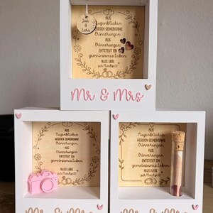 May include: Three white wooden shadow boxes with the text Mr & Mrs in pink. Each box contains a light wood panel with German text and floral engravings. One box features a hanging wooden disc, one a pink camera, and one a glass vial with a cork.