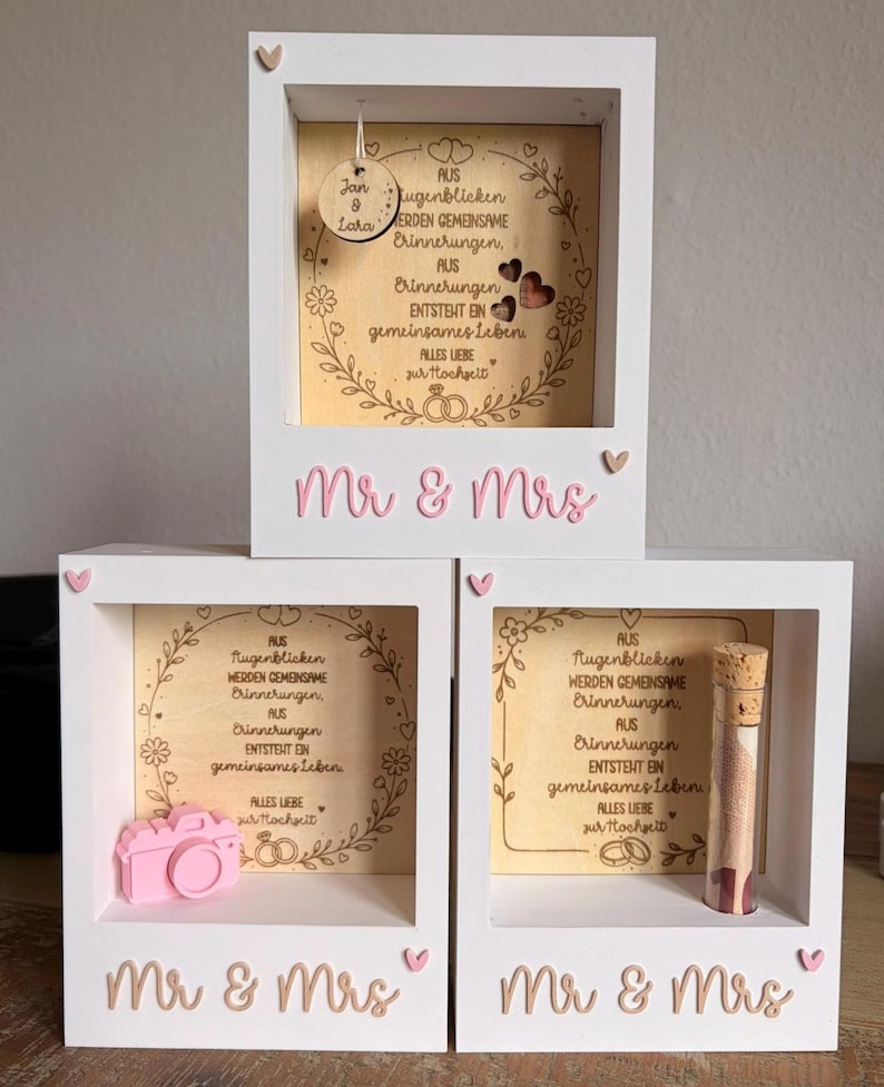 May include: Three white wooden shadow boxes with the text Mr & Mrs in pink. Each box contains a light wood panel with German text and floral engravings. One box features a hanging wooden disc, one a pink camera, and one a glass vial with a cork.