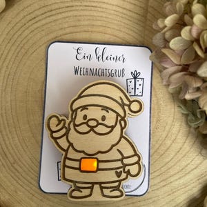 May include: Wooden Santa Claus ornament with an orange light-up belt buckle, mounted on a card with German text. The ornament is a light brown color and is set against a wooden background with decorative beads and dried flowers.