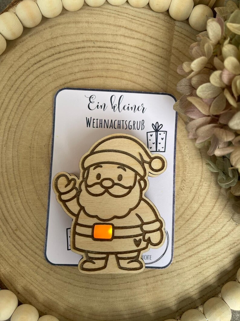 May include: Wooden Santa Claus ornament with an orange light-up belt buckle, mounted on a card with German text. The ornament is a light brown color and is set against a wooden background with decorative beads and dried flowers.