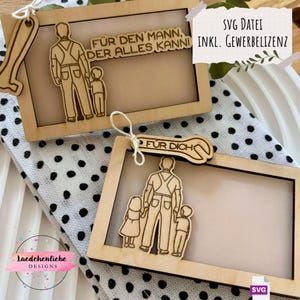 May include: Two wooden picture frames with cutout figures of a parent and children. One frame has the text "FÜR DEN MANN, DER ALLES KANNI" and the other has "FÜR DICH". The frames are on a polka dot fabric.