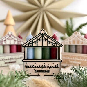 May include: A wooden advent calendar shaped like a Christmas market stall, with four candles in blue, green, and red. The stall has the text "Weihnachtsmarkt ZUM MITNEHMEN". A large paper star and other decorative elements are in the background.