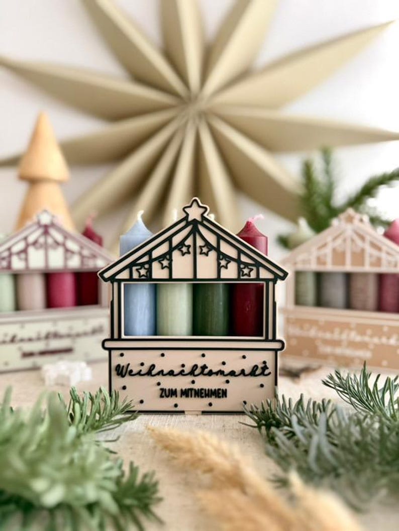 May include: A wooden advent calendar shaped like a Christmas market stall, with four candles in blue, green, and red. The stall has the text "Weihnachtsmarkt ZUM MITNEHMEN". A large paper star and other decorative elements are in the background.