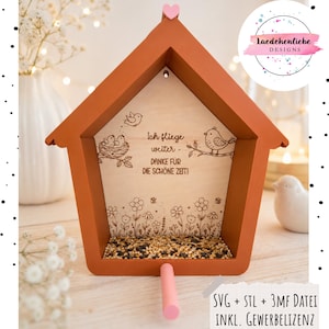 Puede incluir: Un marco de madera con forma de casa, exterior naranja quemado y panel trasero de madera clara. El panel presenta ilustraciones grabadas de pájaros, un nido y flores, con el texto en alemán Ich fliege weiter - DANKE FÜR DIE SCHÖNE ZEIT. Un pequeño corazón rosa corona el techo y un pasador rosa sobresale de la base.